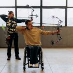 Two archers practice indoors, one using a wheelchair, showcasing determination and skill.